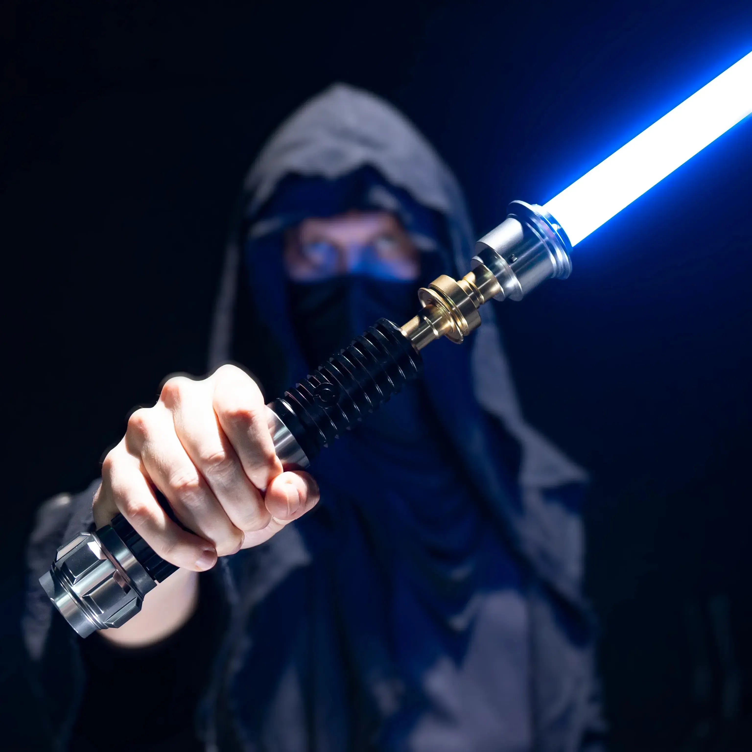 Person in a hooded cloak holding a glowing blue duel-ready aluminum Obi-Wan lightsaber against a black background.