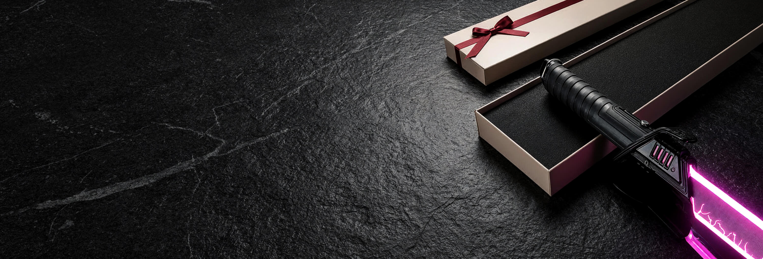 A black lightsaber toy with a glowing pink blade resting in an open presentation box, with a closed gift box tied by a red ribbon on a dark textured background.