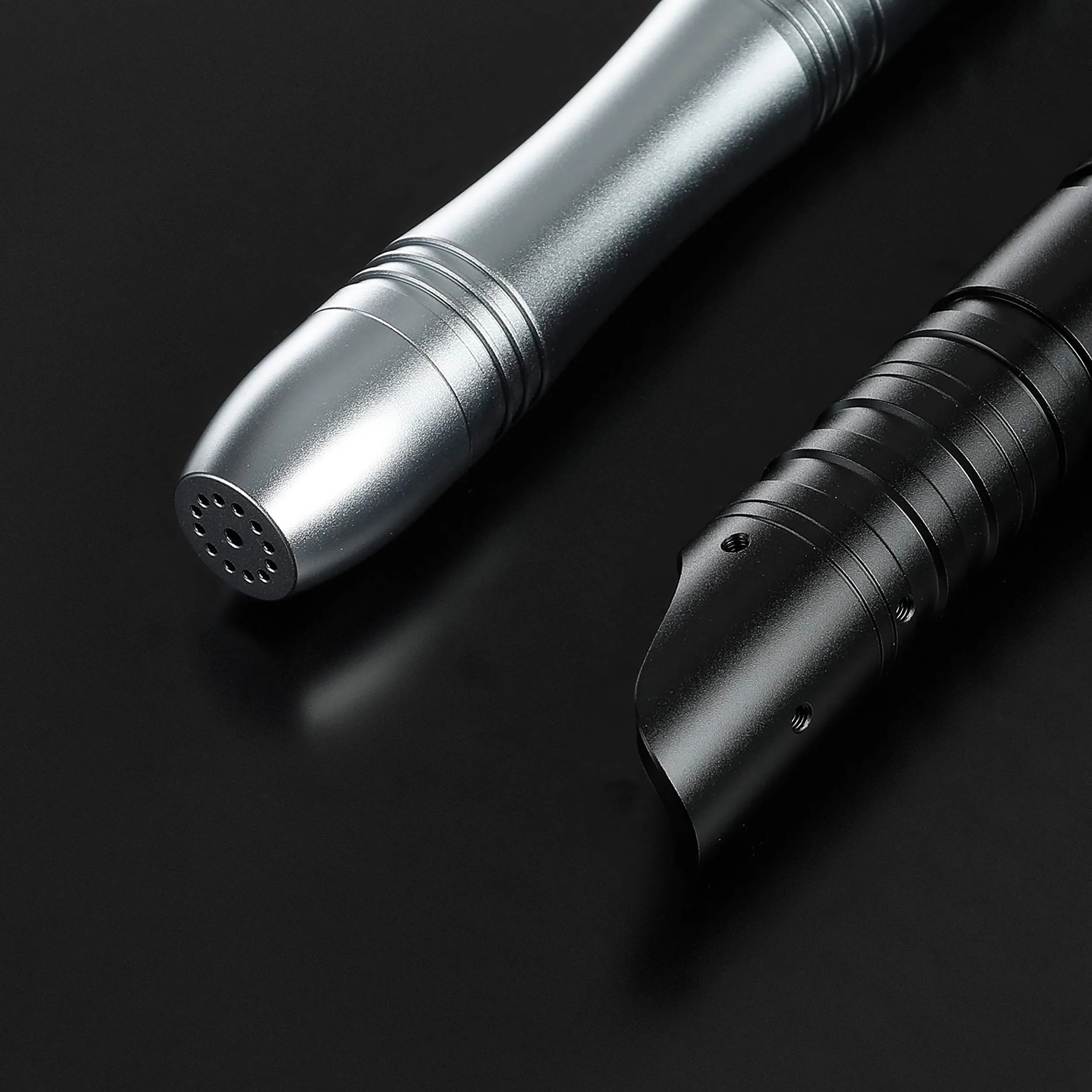 Close-up of two aluminum saber hilts: one silver with a rounded end, one black with a curved emitter.