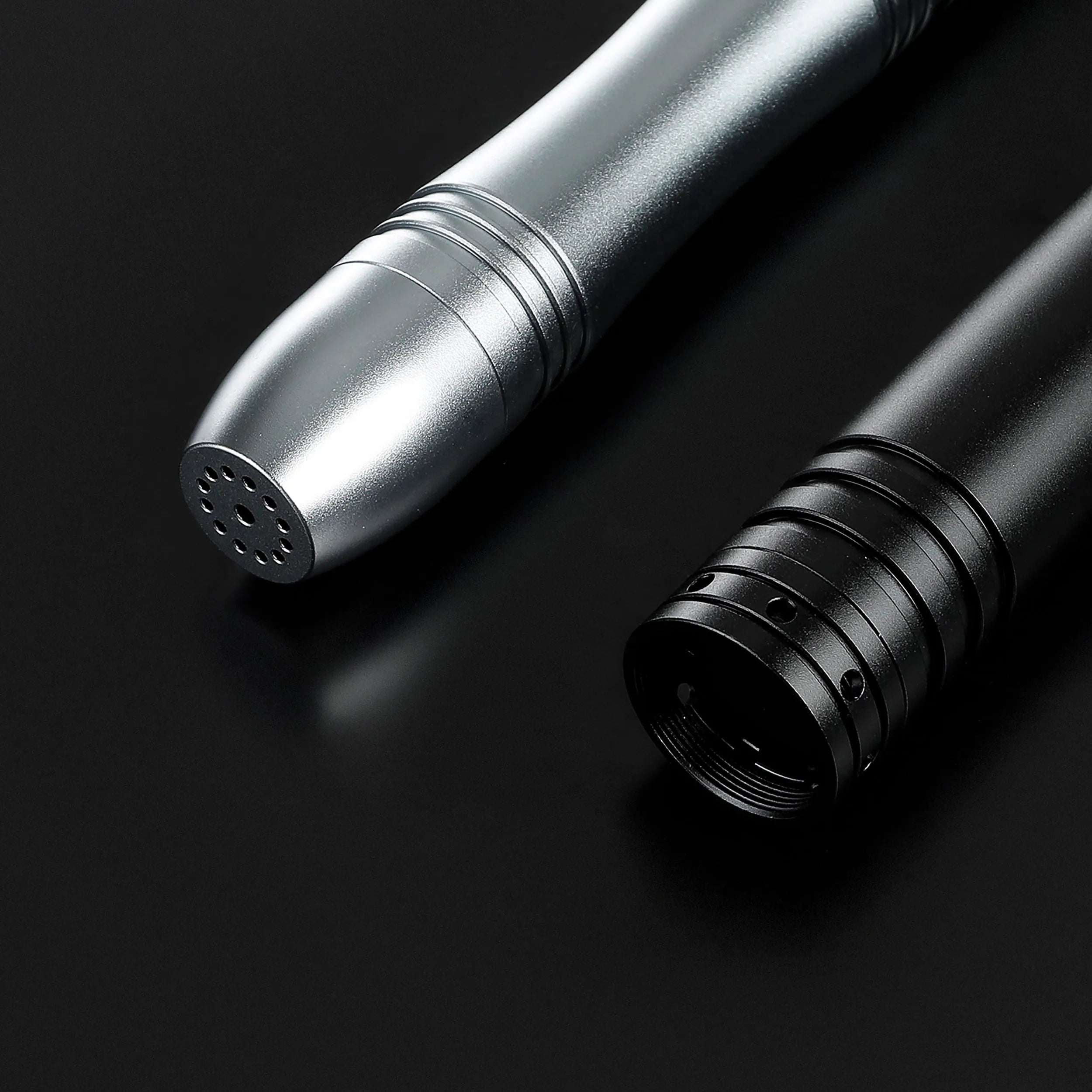 Close-up of two durable aluminum saber hilts, one silver and one black, on a dark background.