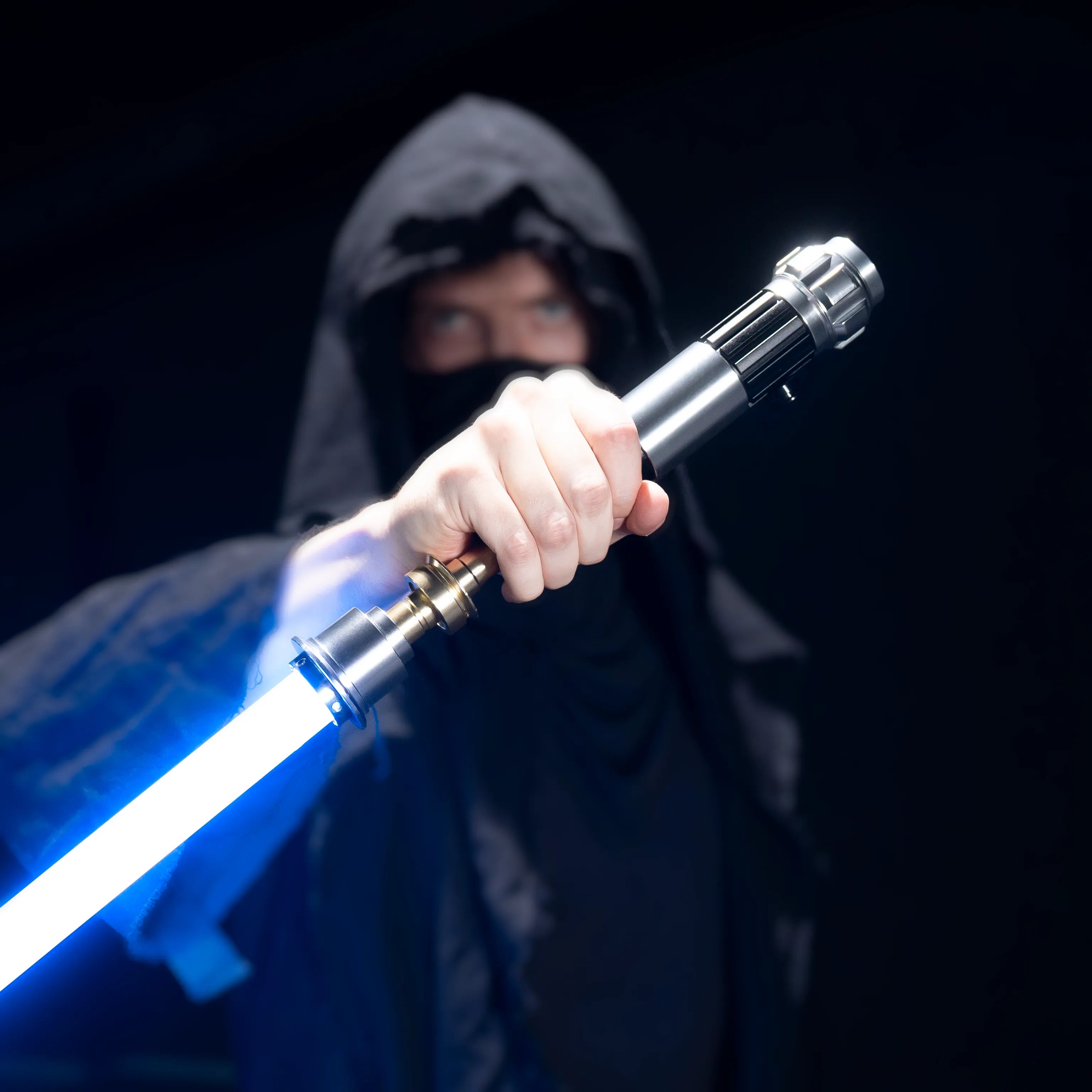 Person in a dark hooded cloak holding a glowing blue T6 aluminum alloy Obi-Wan lightsaber with a duel-ready polycarbonate blade.